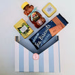 Load image into Gallery viewer, Assorted snacks including a jar of honey, a packet of tea biscuits, and a box of tea leaves on a striped stand.

