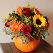 Load image into Gallery viewer, A vibrant autumn floral arrangement featuring orange gerbera daisies, red roses, yellow sunflowers, and green foliage, presented inside a carved orange pumpkin vase on a dark wooden table against a neutral beige wall.
