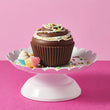 Load image into Gallery viewer, A chocolate cupcake with white frosting and colorful sprinkles displayed on a white scalloped cake stand against a pink background.
