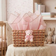 Load image into Gallery viewer, A woven wicker gift basket filled with light pink tissue paper and decorated with a small pink teddy bear, displayed in a soft, nursery-style room setting.
