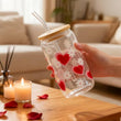Load image into Gallery viewer, Clear tumbler with red heart and white flower designs held by a hand in a cozy living room.
