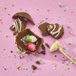 Load image into Gallery viewer, Chocolate Cupcake broken open to reveal colorful gummy candies inside, styled on a pink background with scattered sprinkles and a small wooden toy hammer.
